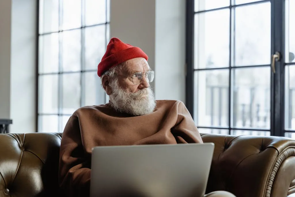 An elderly man in a red beanie is using a laptop indoors, sitting comfortably on a couch.